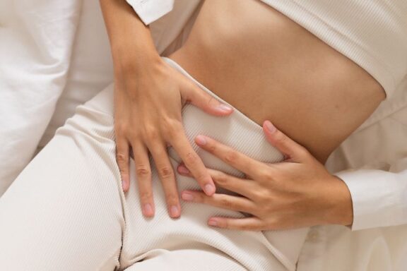 Ujęcie tułowia leżącej na łóżku kobiety, która trzyma się za brzuch w geście bólu.