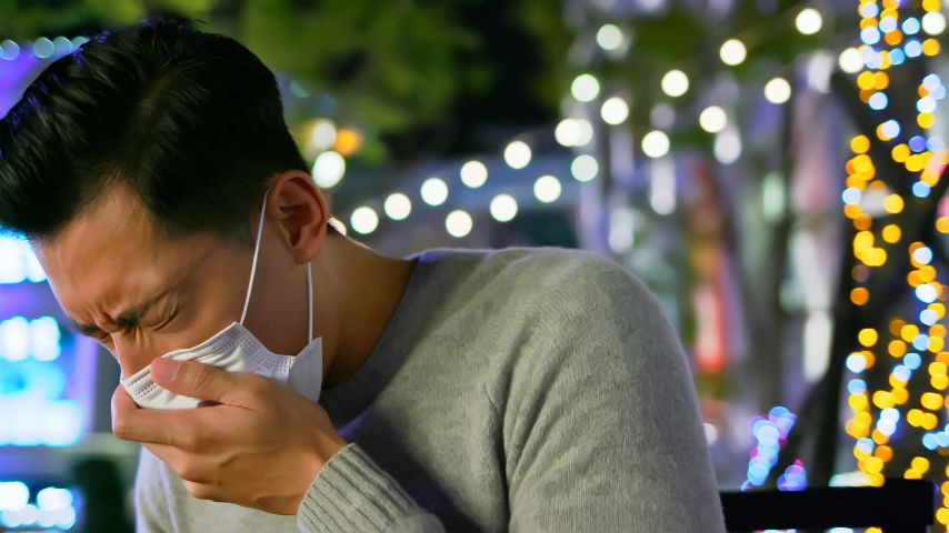 Kaszlący mężczyzna w maseczce, który przytrzymuje dłońmi twarz.