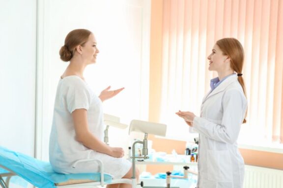 Pacjentka siedząca na kozetce prowadzi rozmowę z ginekolożką stojącą naprzeciw.